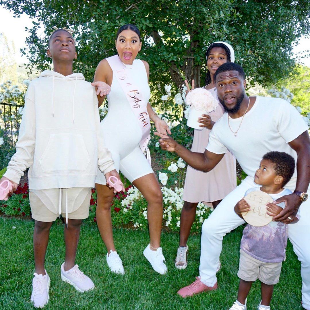 Eniko Hart poses with family members in matching white outfits on a lawn with ivy-covered wall