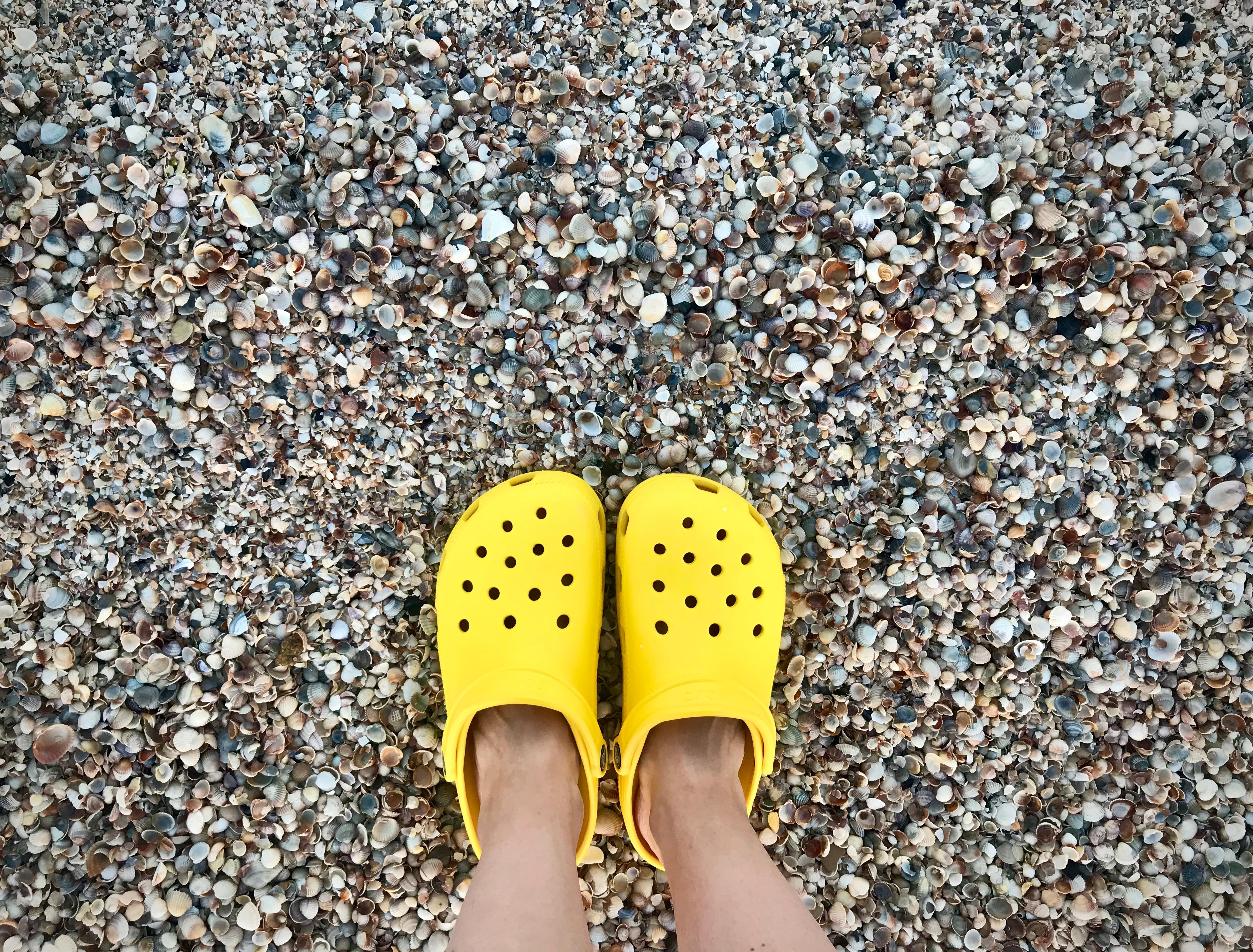 Bright yellow Crocs clogs with ventilation holes worn on a beach covered in seashells