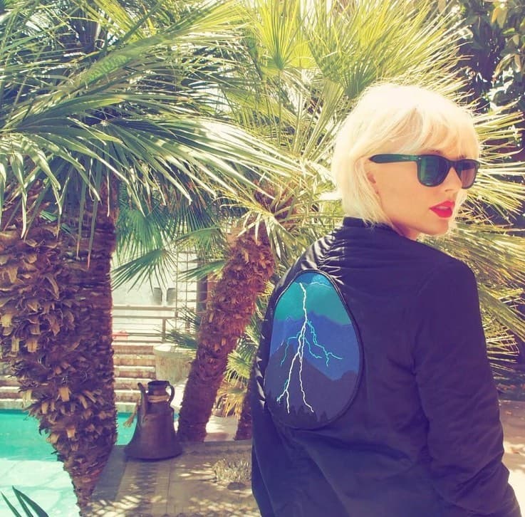 Person wearing sunglasses and black jacket with lightning bolt design, standing among palm trees