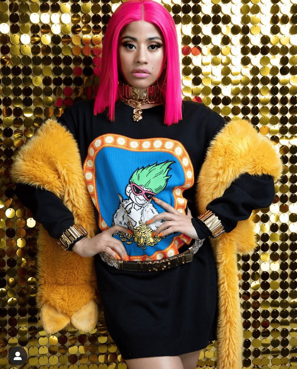 Hennessy Carolina wearing bright pink hair, black graphic tee, and yellow fur stole against gold