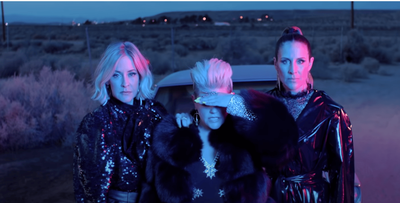 Four women in sparkly black outfits standing in a desert landscape at dusk