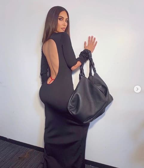 Woman in black backless dress holding black handbag against white wall