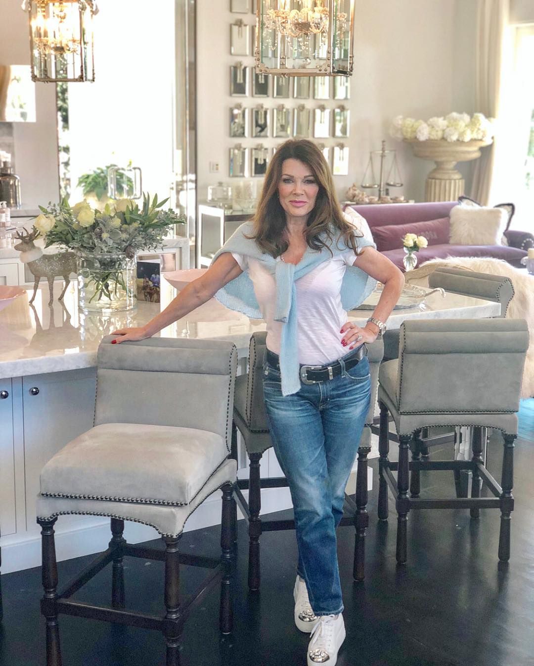 Lisa Vanderpump poses in her elegant living room with gray upholstered chairs and white flowers.
