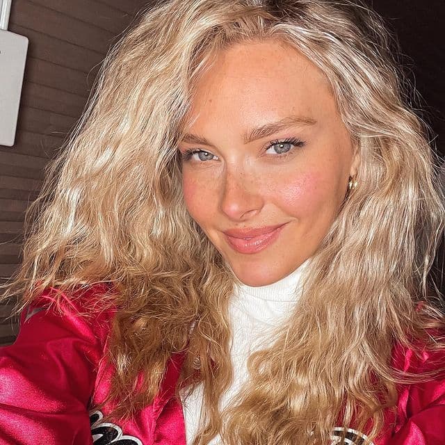 Camille Kostek smiles at the camera wearing a red jacket and white turtleneck