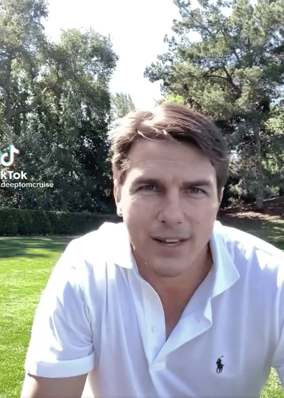Man in white polo shirt speaking to camera outdoors with trees and lawn behind him