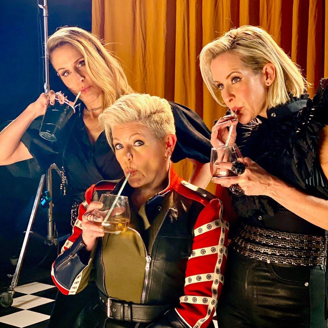 Three women drinking together at an indoor event with gold curtains in the background