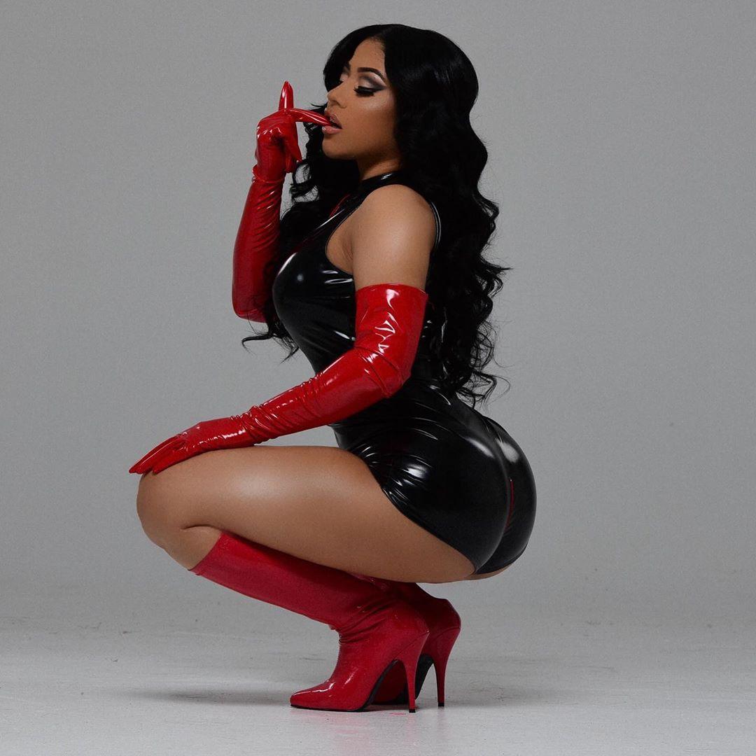Woman in black latex dress and red patent leather gloves and boots against gray background