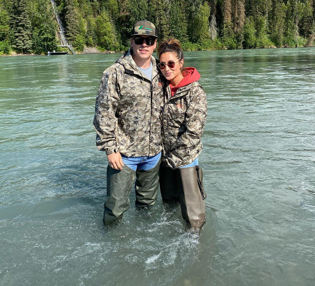 Two people in waders standing in a turquoise river with forested banks